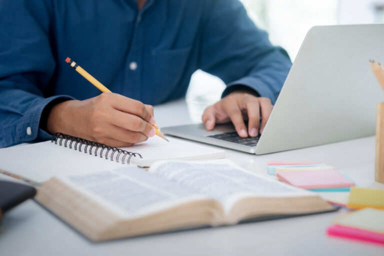 Personne prenant des notes dans un carnet tout en travaillant sur un ordinateur portable, illustrant la méthode de travail hybride de l’Union des Scribes